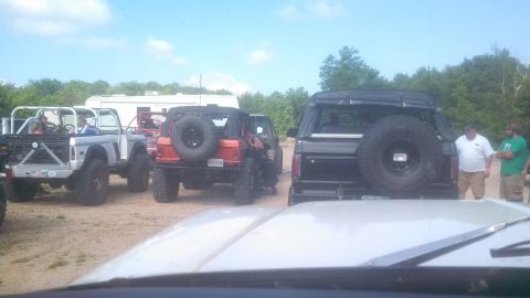 1979 Ford Bronco - Several off-road vehicles, including a gray Ford Bronco with a roll cage and a black Bronco with a large spare tire, are parked on a dirt l…