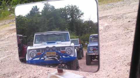 1979 Ford Bronco - A reflection in a side mirror shows a line of Ford Broncos driving on a dirt road. The closest Bronco is silver with a blue bumper and has…