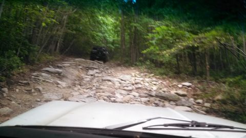 1979 Ford Bronco - View from inside a vehicle driving up a rocky, wooded trail. A black SUV is visible ahead on the trail.