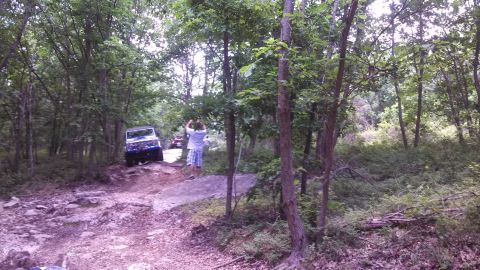 1979 Ford Bronco - A blue SUV is navigating a rocky, wooded trail. A person stands next to the trail, facing away from the camera.