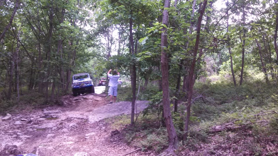 Blue SUV on a rocky trail in a forest with a person standing nearby.