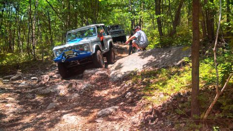 1979 Ford Bronco - A silver 1979 Ford Bronco with a blue grille guard is navigating a rocky, wooded trail. Another off-road vehicle is visible behind it, and…