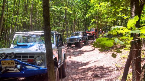 1979 Ford Bronco - Three vintage Ford Broncos drive in single file on a rocky, wooded trail. The lead Bronco is white with a blue accent, followed by a blue B…