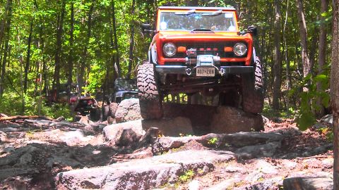 1979 Ford Bronco - An orange Ford Bronco is navigating a rocky trail in a wooded area. The vehicle is lifted and has large off-road tires, with a winch mounte…