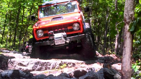 1979 Ford Bronco - An orange Ford Bronco is navigating a rocky trail in a forest. The vehicle is angled towards the camera, showing its front grille, winch, a…