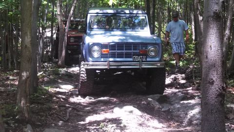 1979 Ford Bronco - A light blue Ford Bronco is shown driving on a rocky, wooded trail. A man in a t-shirt and plaid shorts is walking behind the Bronco.