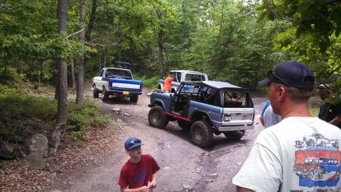 1979 Ford Bronco - A group of off-road vehicles, including a blue Ford Bronco and a white Ford pickup truck, are parked on a dirt trail in a wooded area.