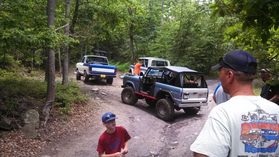 Off-road vehicles including a blue Ford Bronco and a white Ford pickup truck on a dirt trail in a forest.