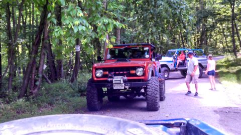 1979 Ford Bronco - A bright red Ford Bronco is parked on a dirt road in a wooded area, with other vehicles and people visible in the background. The Bronco ha…
