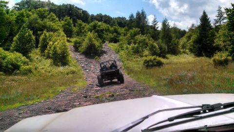 1979 Ford Bronco - A black off-road buggy drives up a rocky, dirt path through a wooded area. The photo is taken from the perspective of another vehicle, show…