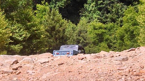 1979 Ford Bronco - A blue first-generation Ford Bronco is partially submerged in loose dirt and rocks, with a dense forest in the background.