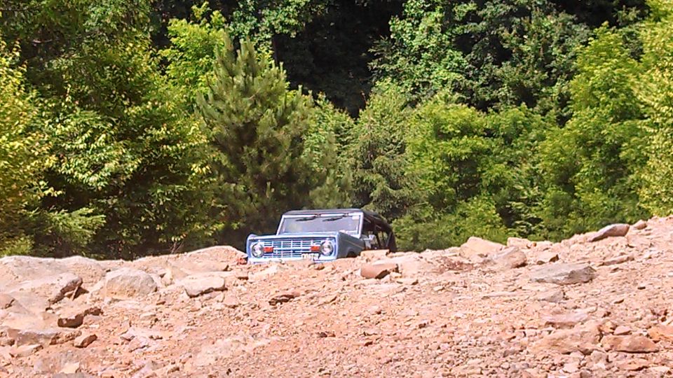 Blue first-generation Ford Bronco stuck in dirt and rocks with trees behind it.