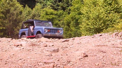 1979 Ford Bronco - A blue 1979 Ford Bronco is navigating a rocky, off-road trail with dense green trees in the background. The vehicle has its top removed, re…