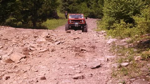 1979 Ford Bronco - An orange Ford Bronco is driving up a rocky, off-road trail. The vehicle is viewed from the front, showing its large tires and winch.