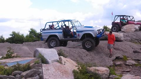 1979 Ford Bronco - A silver and blue off-road SUV with a roll cage is navigating a rocky terrain. A man in an orange shirt stands on the rocks near the front…