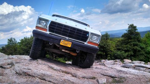 1979 Ford Bronco - A white 1979 Ford Bronco is angled up a rocky incline against a backdrop of lush green trees and a blue sky with dramatic clouds. The front…