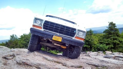 1979 Ford Bronco - A white 1979 Ford Bronco is shown at an extreme angle, navigating a rocky trail with a scenic mountain vista in the background. The front o…