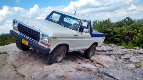 1979 Ford Bronco - A white and blue Ford Bronco is navigating a rocky, off-road terrain with a wooded hillside in the background under a cloudy sky.