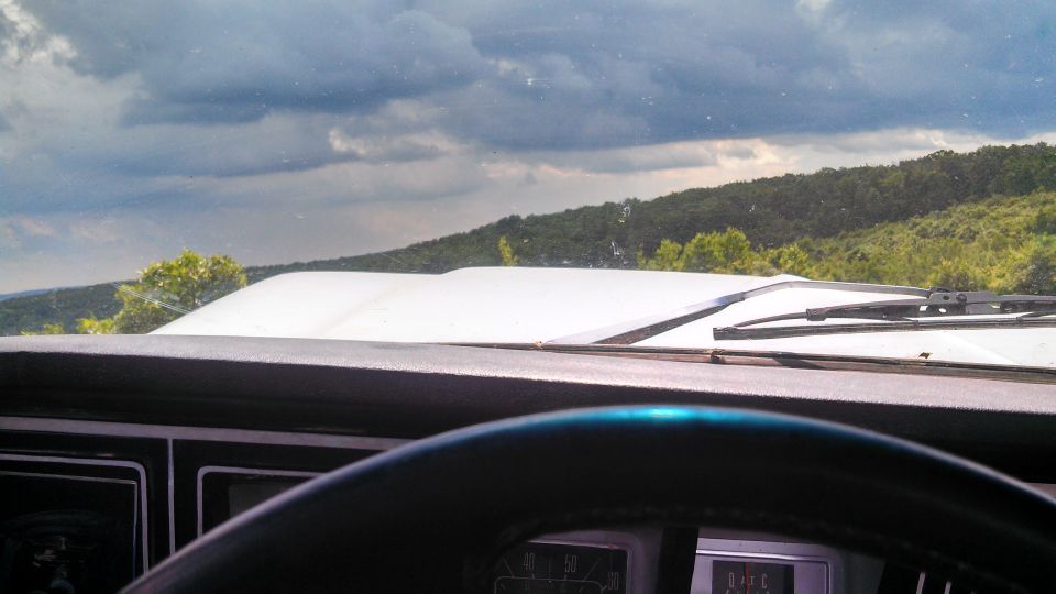 Interior view of a 1979 Ford Bronco dashboard and hood, looking out at a cloudy sky and wooded hills.