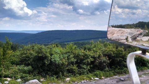 1979 Ford Bronco - A view from a vehicle, likely a 1979 Ford Bronco, looking out over a forested mountain landscape. A side mirror reflects a large rock forma…