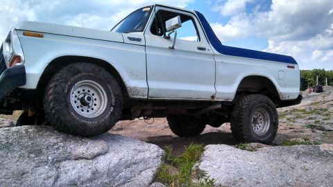1979 Ford Bronco - A white 1979 Ford Bronco with a blue top is shown from the side, navigating a rocky terrain. The vehicle's large off-road tires are clearly…