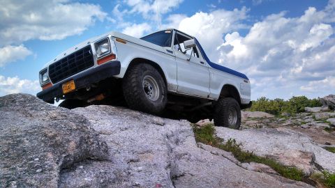 1979 Ford Bronco - A white 1979 Ford Bronco with a blue top is parked on a rocky terrain under a cloudy blue sky. The vehicle is angled upwards, with its fron…