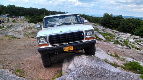 1979 Ford Bronco - A white 1979 Ford Bronco is shown from the front, driving over large rocks on a rocky, wooded trail. The vehicle has a Virginia license pla…