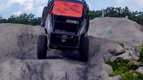 1979 Ford Bronco - The rear view of a custom off-road vehicle, possibly a modified Toyota, is shown climbing a rocky incline. A sign on the back reads "BE PRE…
