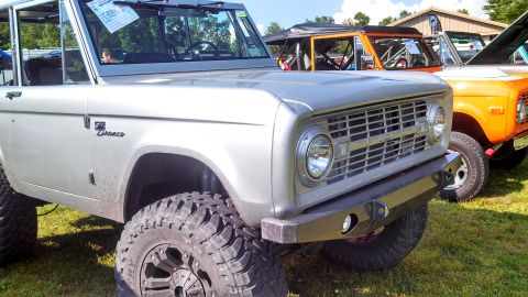 1979 Ford Bronco - A silver first-generation Ford Bronco is parked on grass, with the front end and driver's side visible. An orange first-generation Ford Bro…