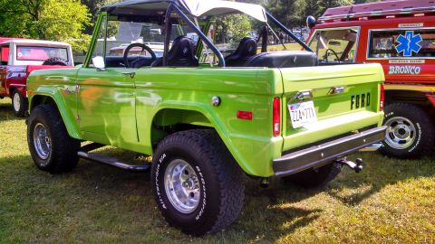 1979 Ford Bronco - The rear quarter view of a bright green 1979 Ford Bronco is shown parked on a grassy area. The vehicle has a black soft top, chrome side st…