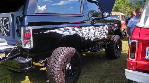 1979 Ford Bronco - The rear quarter panel of a black lifted Ford Bronco is shown with a skull and flag graphic. A large off-road tire and chrome exhaust tip a…
