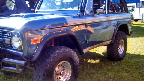 1979 Ford Bronco - The right side of a dark blue 1979 Ford Bronco is shown parked on grass. The vehicle has large off-road tires, a chrome bumper, and a silve…