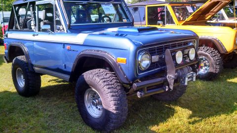 1979 Ford Bronco - A blue 1979 Ford Bronco with a white stripe is parked on grass next to a yellow vehicle. The blue Bronco has large off-road tires, a winch,…