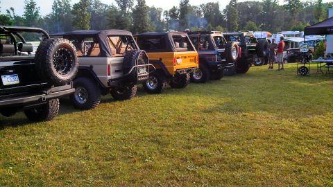 1979 Ford Bronco - A line of Ford Broncos are parked on a grassy field with trees in the background. The vehicles are modified with large tires and off-road a…