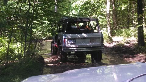 1979 Ford Bronco - A lifted, silver 1979 Ford Bronco with a soft top is driving through a shallow stream in a wooded area. The rear of the vehicle is visible,…