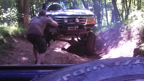 1979 Ford Bronco - A person pushes on the rear of a dark-colored SUV that is stuck on a dirt incline in a wooded area. The front of the SUV, with its winch an…