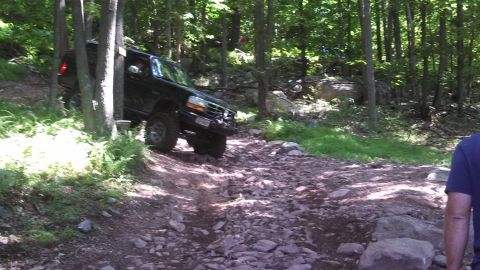 1979 Ford Bronco - A black Ford Bronco is navigating a rocky, wooded trail. The vehicle is angled uphill, with its front wheels turned to the right.