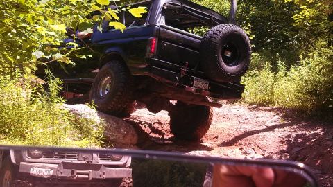 1979 Ford Bronco - A black Ford Bronco is navigating a muddy, rocky trail with dense green foliage on either side. The vehicle is angled, showing its rear tir…
