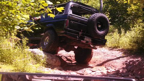 1979 Ford Bronco - A black 1979 Ford Bronco is shown from the rear-driver side, navigating an off-road trail with large tires and a lifted suspension. The veh…