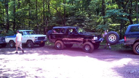 1979 Ford Bronco - A white Ford pickup truck and a black Ford Bronco are parked on a dirt road in a wooded area. The Bronco is being towed by another vehicle,…