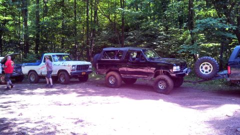 1979 Ford Bronco - A black Ford Bronco is parked next to a white Ford pickup truck in a wooded area. The Bronco is equipped with large off-road tires and a sp…