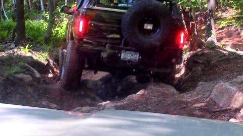 1979 Ford Bronco - The rear of a dark-colored SUV, possibly a Ford Bronco, navigates a rocky, wooded trail. The vehicle's red taillights are illuminated as it…