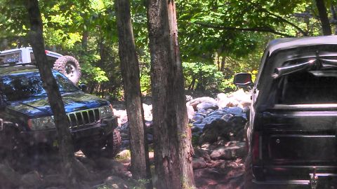 1979 Ford Bronco - Two off-road vehicles are navigating a rocky, wooded trail. The vehicle in the foreground is a dark-colored SUV, possibly a Jeep Grand Cher…