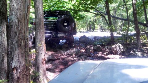 1979 Ford Bronco - A black SUV with a spare tire mounted on the rear is navigating a rocky, wooded trail. The vehicle is positioned between trees and large bo…