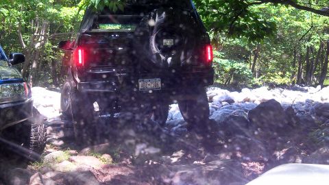 1979 Ford Bronco - The rear view of a black SUV is shown navigating a rocky terrain with a spare tire mounted on the back. The license plate reads "ADZ-2506"…