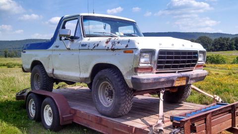 1979 Ford Bronco - A white and blue 1979 Ford Bronco is parked on a wooden trailer, with a grassy field and trees in the background. The truck shows signs of…