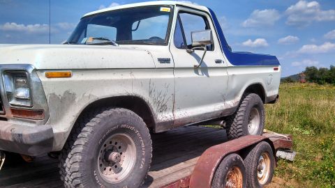 1979 Ford Bronco - A dirty white 1979 Ford Bronco with a blue top is parked on a trailer in a grassy field under a cloudy blue sky. The side of the Bronco sho…