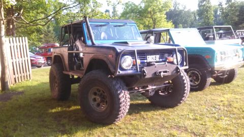 1979 Ford Bronco - A blue 1979 Ford Bronco with a roll cage and large off-road tires is parked on a grassy lawn next to a teal Bronco. The front of the blue B…