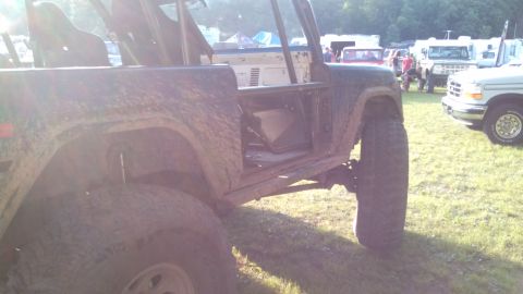 1979 Ford Bronco - A muddy, open-door 1979 Ford Bronco sits on a grassy field with other vehicles and tents in the background. The vehicle's interior and roll…