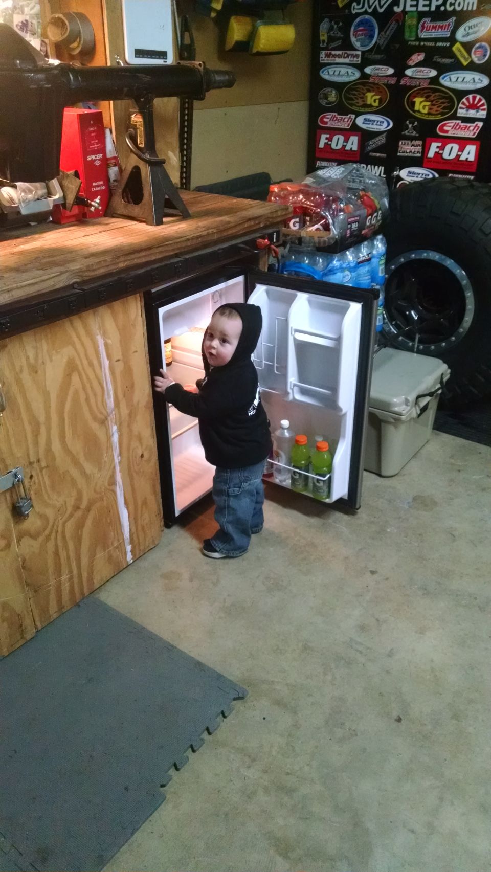 Toddler in a black hoodie and jeans looking into an open mini-fridge in a garage.